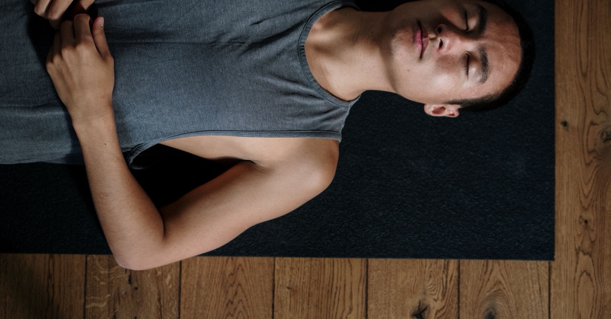 Body Scan Übung Mann liegt entspannt mit geschlossenen Augen und den Händen auf dem Bauch auf einer Matte, Sinnbild für den Body Scan